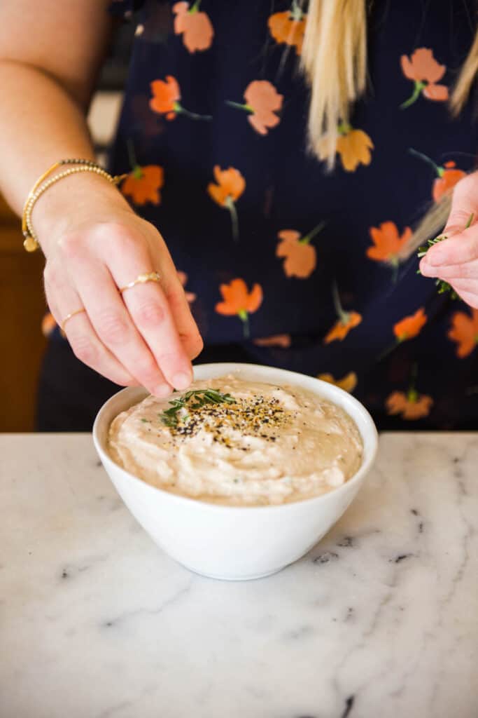 Woman garnishing a dip in a white bowl with fresh roseamary.