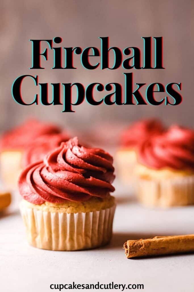 Red Frosting on top of a Fireball flavored cupcake with a cinnamon stick on the table in front of it and more cupcakes behind it.