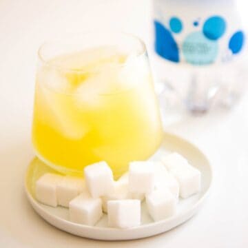 Sugar cubes on a small white plate along with a cocktail and a bottle of club soda behind it.