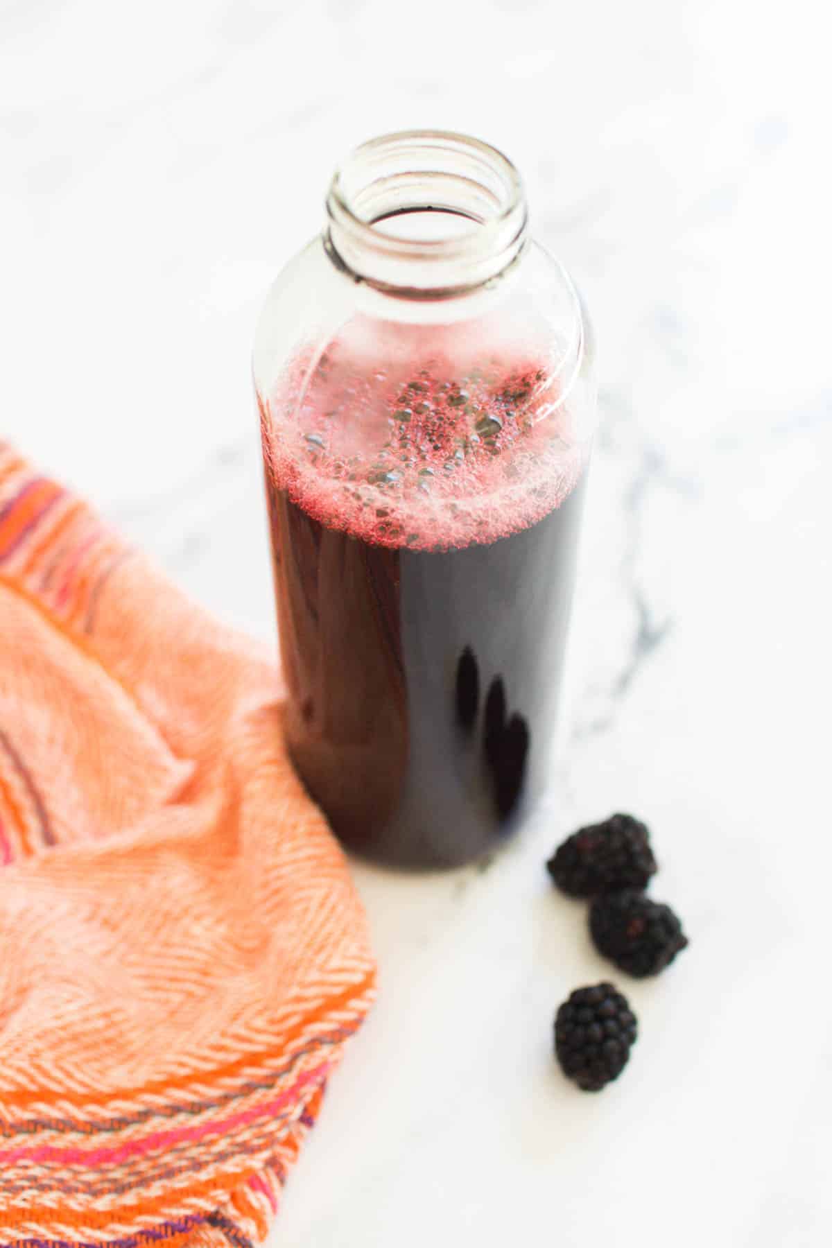 Bottle of blackberry cocktail syrup on white counter with fresh blackberries laying nearby.