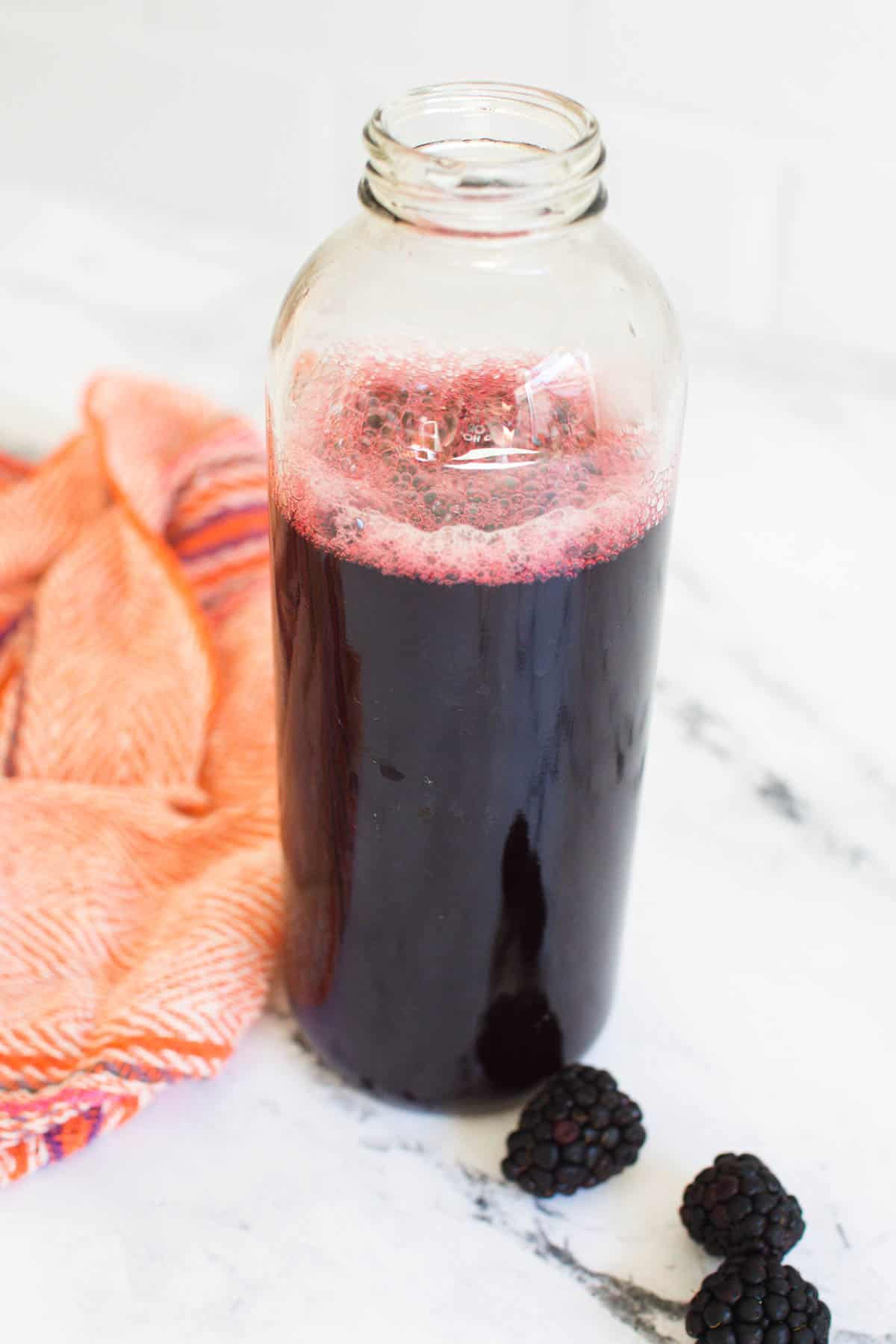 Blackberry cocktail syrup in clear jar on a counter next to fresh berries and a cloth napkin.  