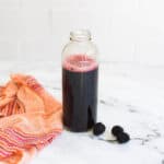 Blackberry cocktail syrup in jar with towel and fresh blackberries on orange counter.