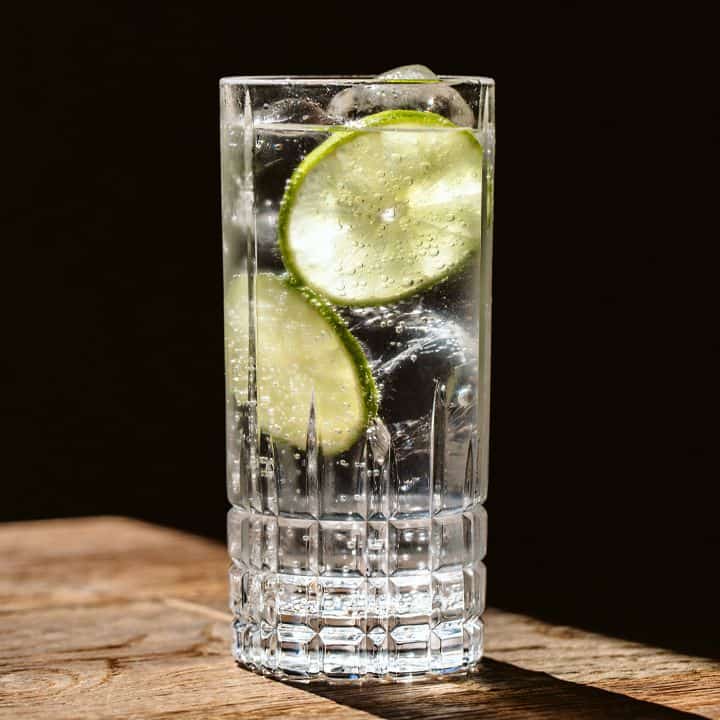 Close up of a gin and tonic with lime slices floating in a tall glass on a wood table.