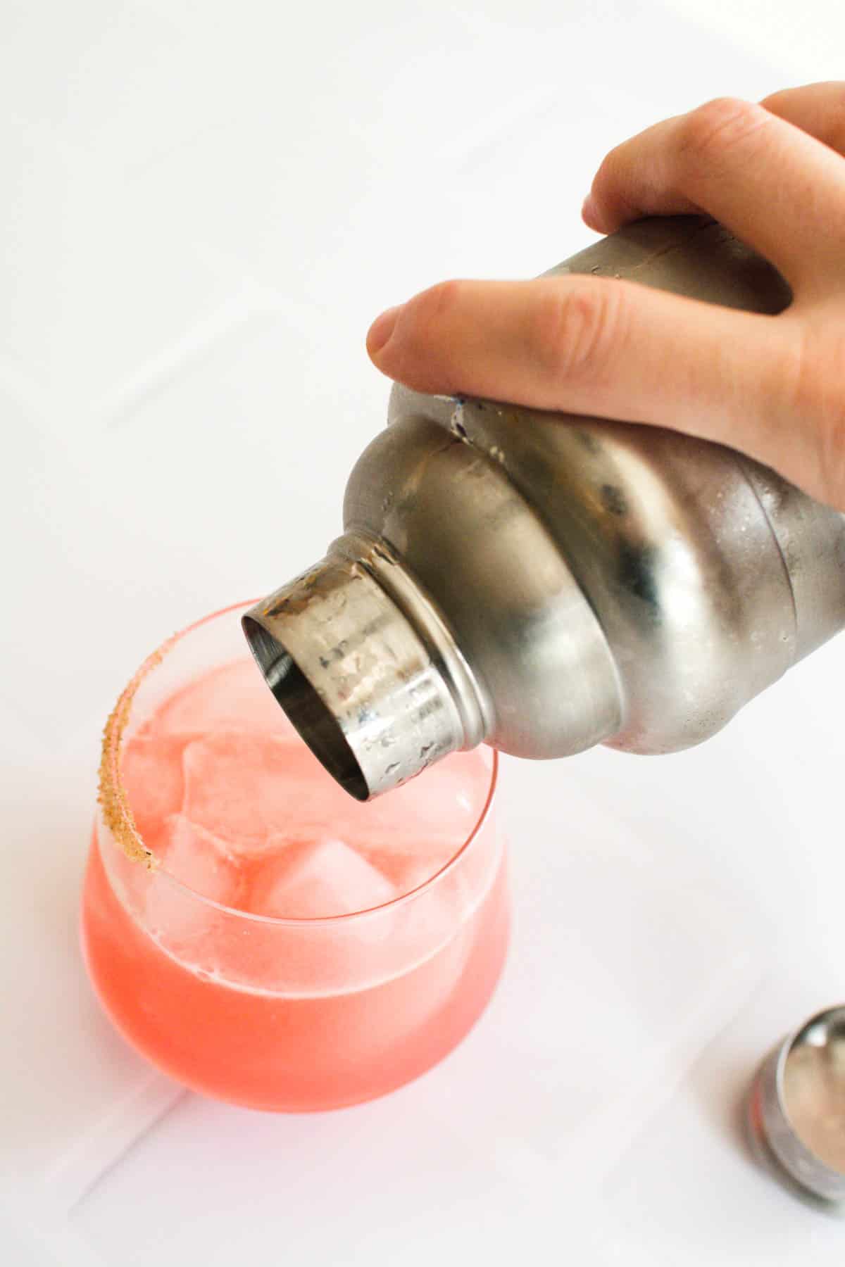 A hand pouring blood orange margarita out of a cocktail shaker into a cocktail glass.