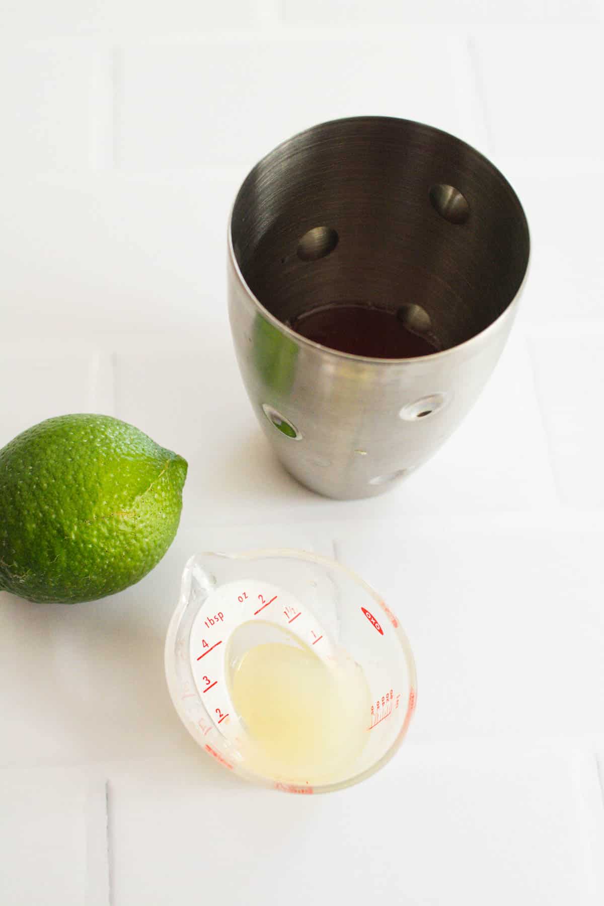 Fresh lime juice in a measuring cup next to a cocktail shaker.