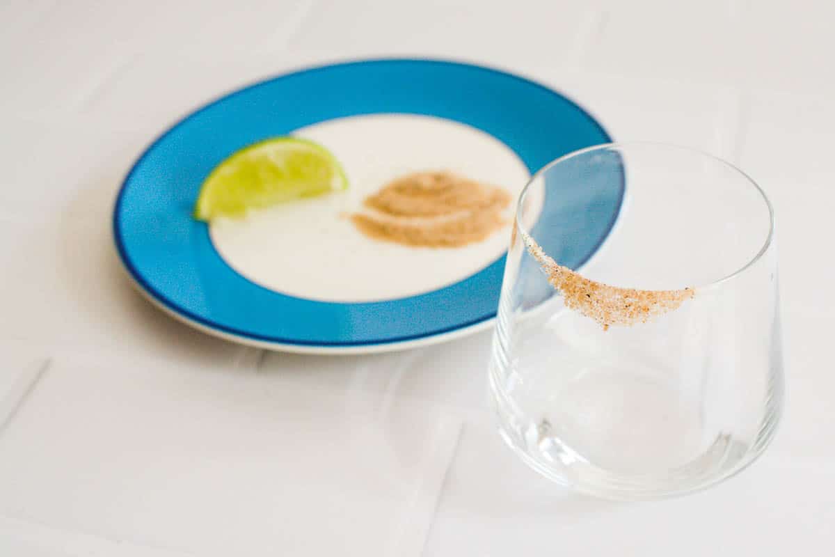 A plate with a lime wedge and spiced sugar on a plate next to a cocktail glass with a sugar rim.