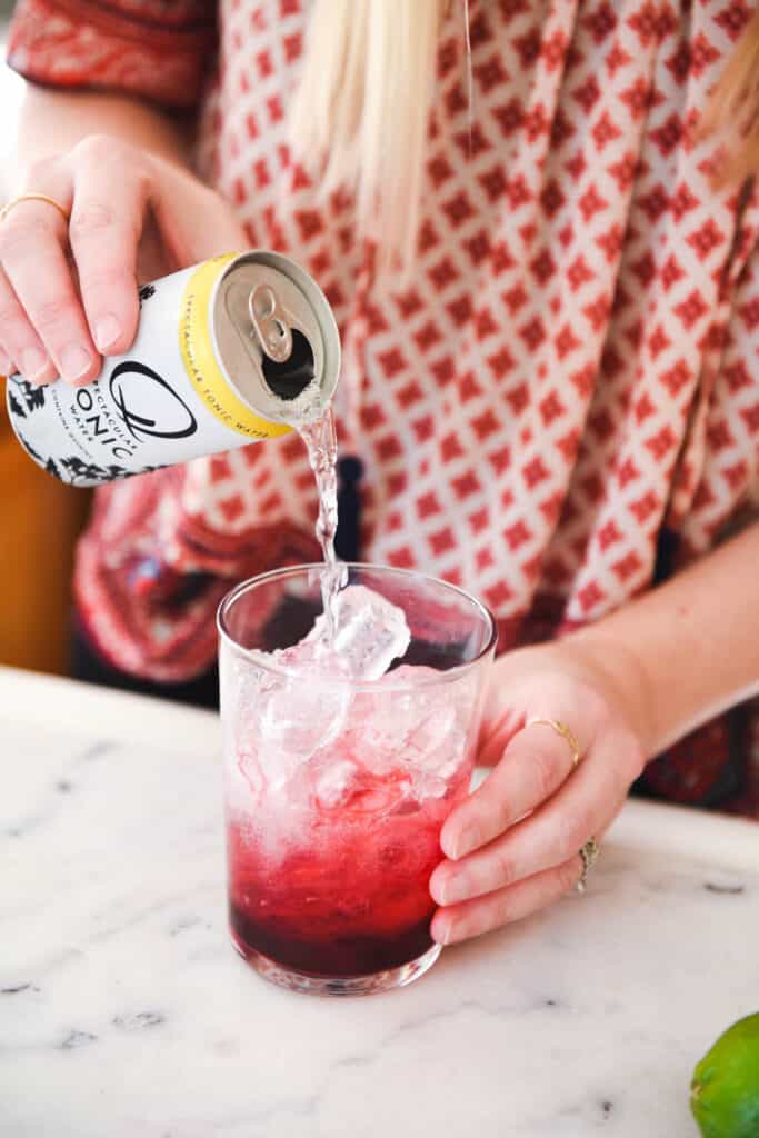 Woman pouring tonic in a cocktail glass full of ice and blackberry simple syrup.