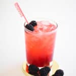 Close up of a glass full of blackberry gin and tonic with fresh blackberries as a garnish and a fun name straw, sitting on a gold coaster.