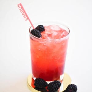 Close up of a glass full of blackberry gin and tonic with fresh blackberries as a garnish and a fun name straw, sitting on a gold coaster.