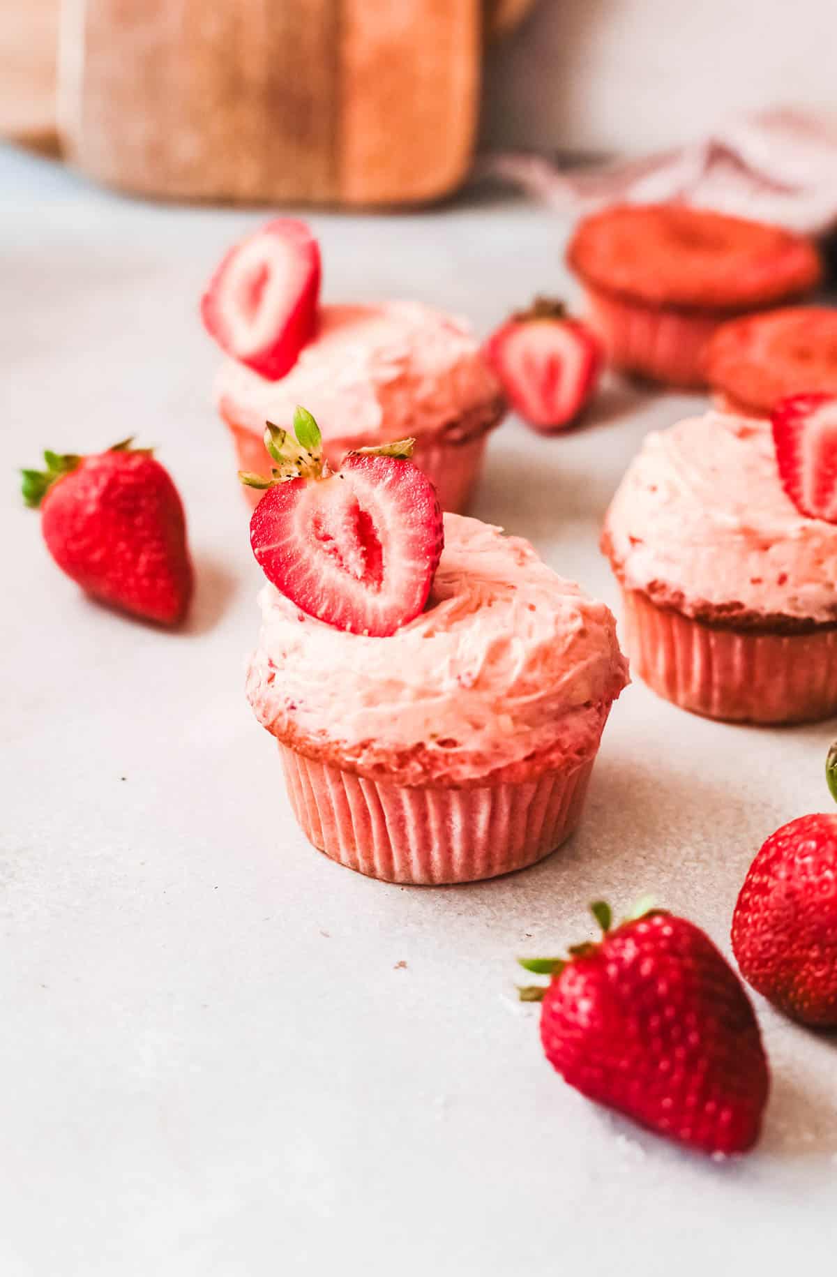 Close up of a frosted Strawberry cupcake topped with a sliced strawberry and more cupcakes behind it. 