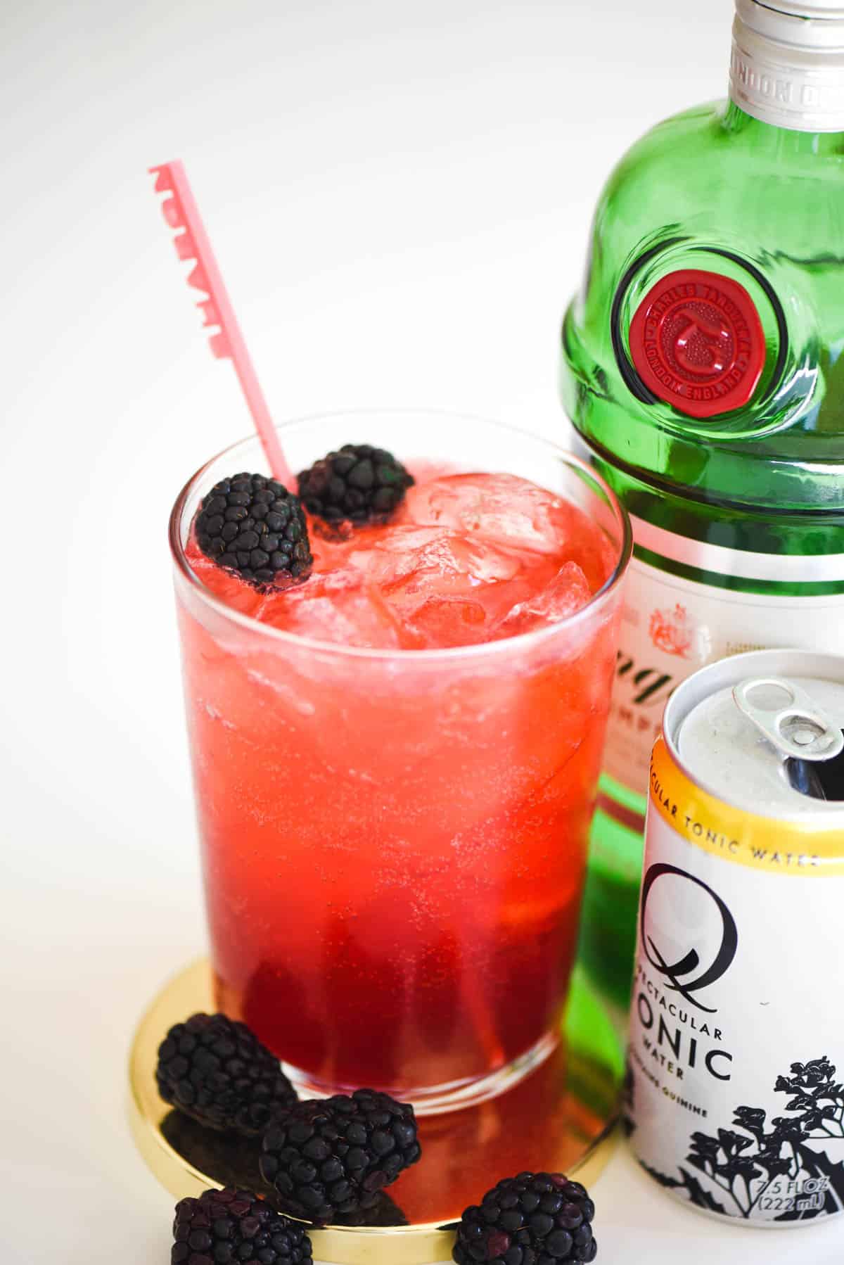 A cocktail glass with a blackberry gin and tonic on the table next to a bottle of gin.