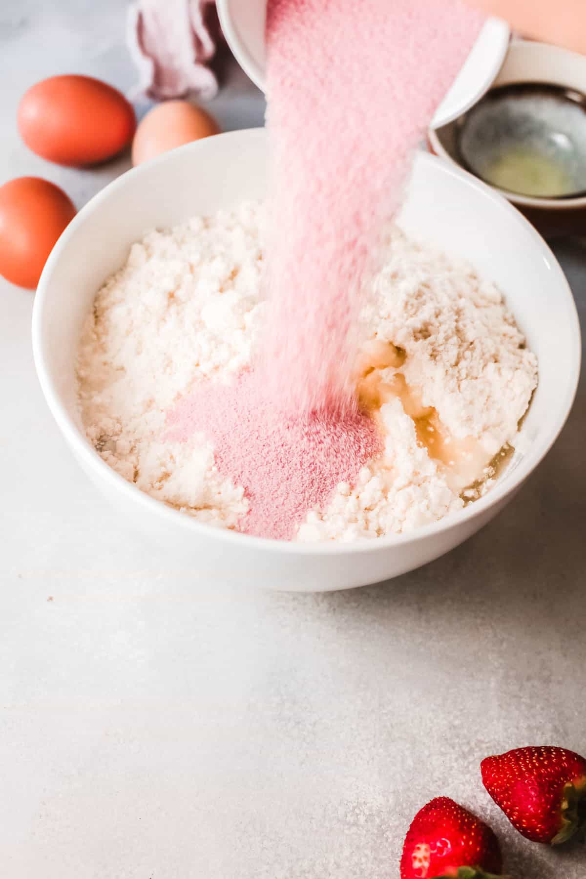 A bowl of dry cake mix on a counter with dry strawberry Jello powder being added. 