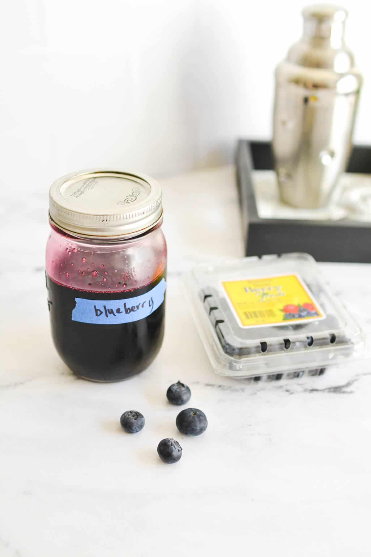 Blueberry Simple Syrup in a jar labeled with a piece of tape next to a container of bluberries. 