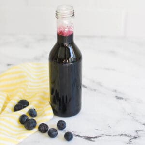 glass container of blueberry syrup surrounded by blueberries on a marble counter