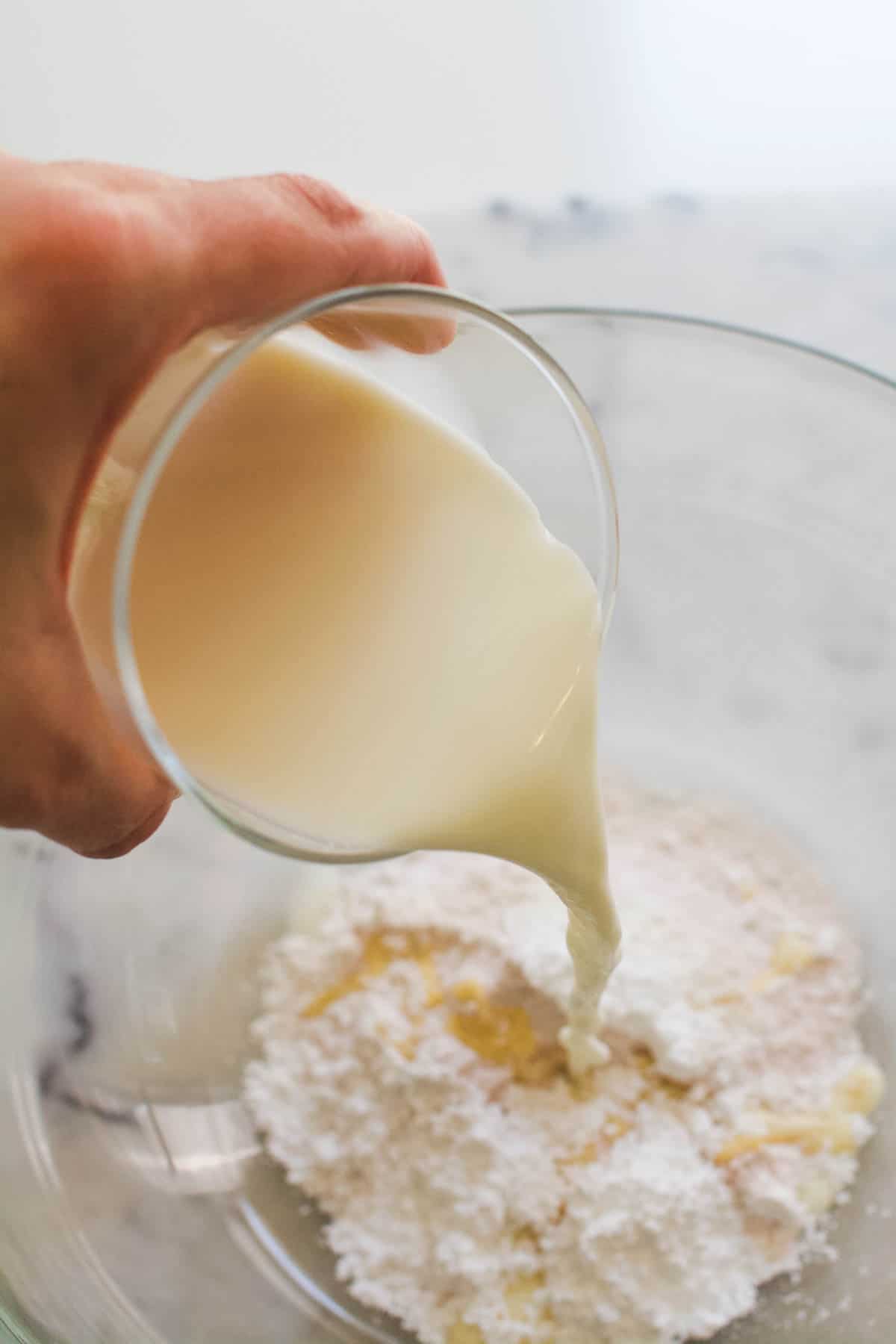 A hand pouring a glass of milk into a glass mixing bowl with powdered vanilla pudding.