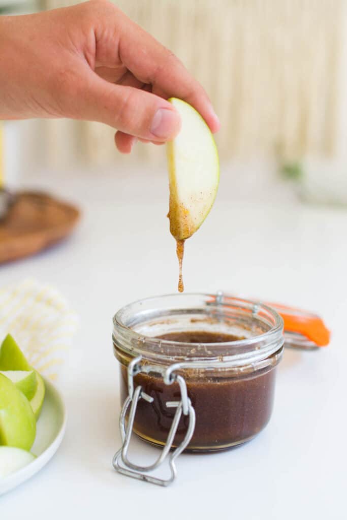 Hand dipping a sliced green apple into a honey dip in a jar.
