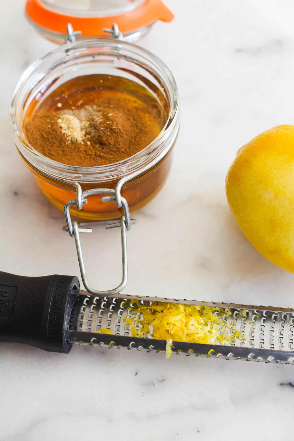 A small jar of honey next to a lemon and a zester tool with lemon zest on it.