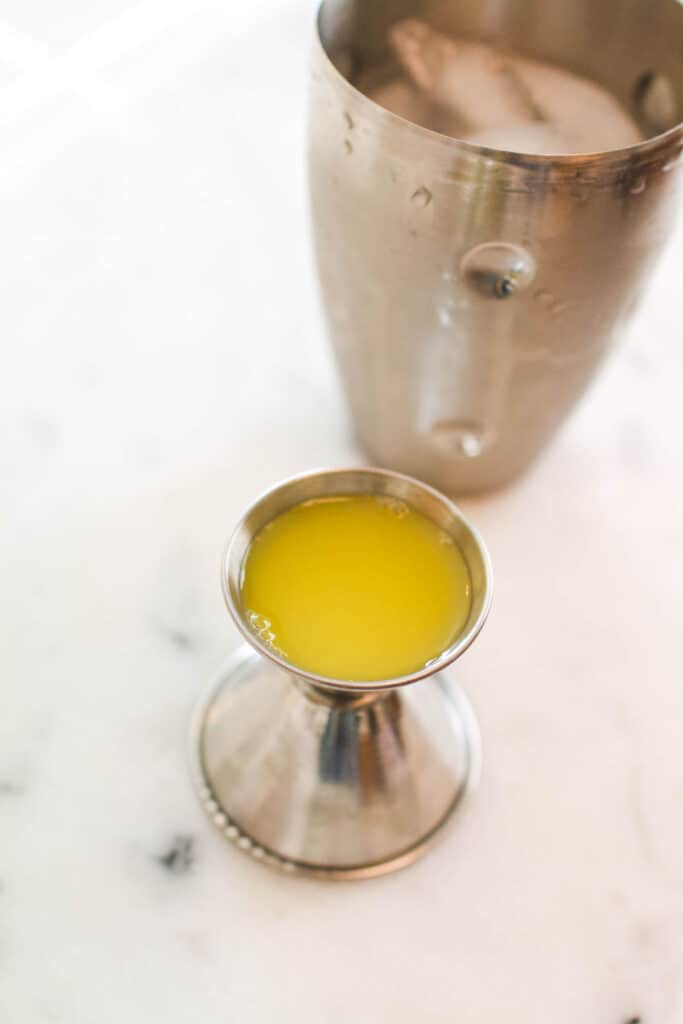 A cocktail shaker on a counter next to a jigger with orange juice.