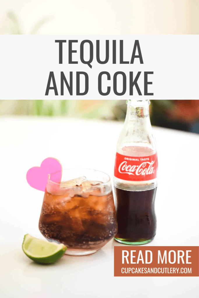 A coke bottle on a table next to a cocktail in a short glass with an acrylic heart glass charm.