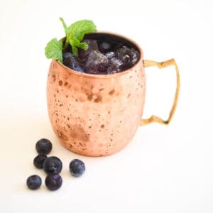 A copper mug full of blueberry moscow mule cocktail with fresh blueberries laying on the counter.