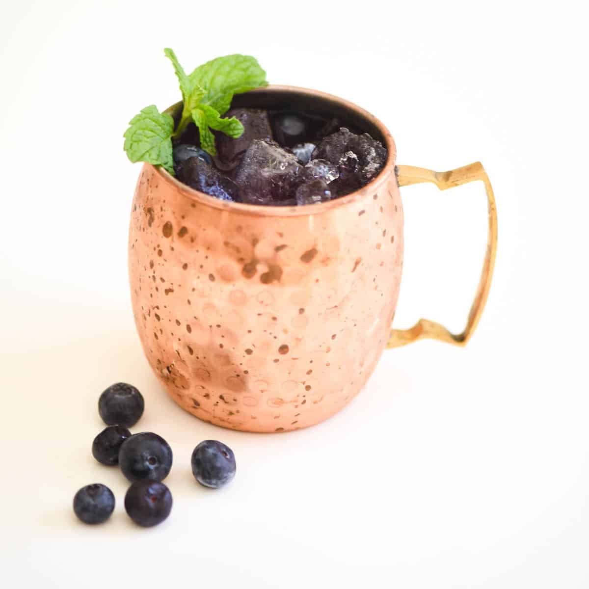 A copper mug full of blueberry mule cocktail with fresh blueberries laying on the counter. 