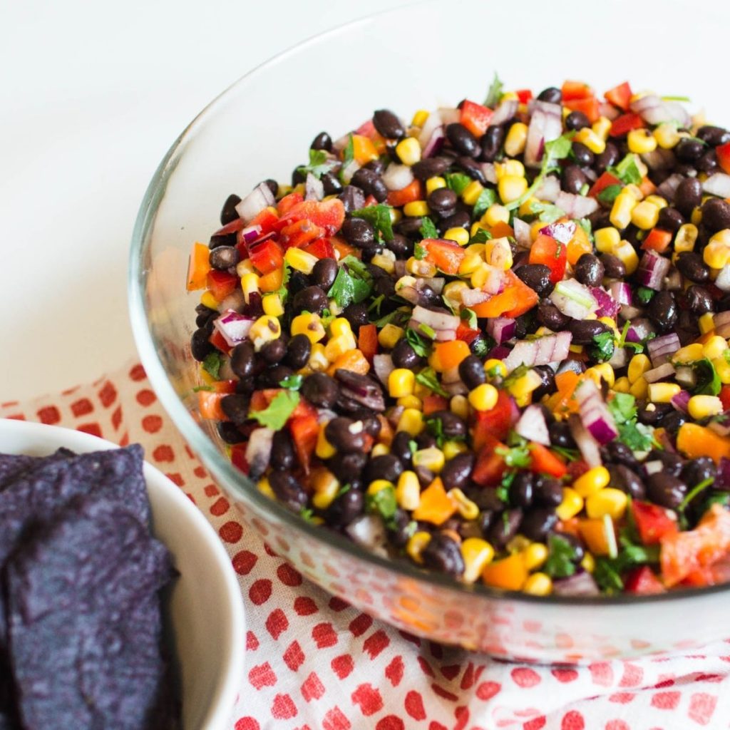 A bowl of black bean salad on a table next to a bowl of blue corn tortilla chips.