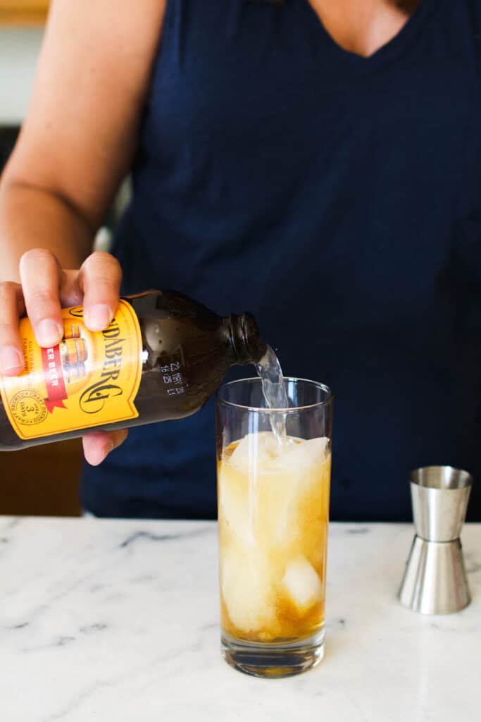 A woman adding ginger beer to a cocktail glass on a counter.