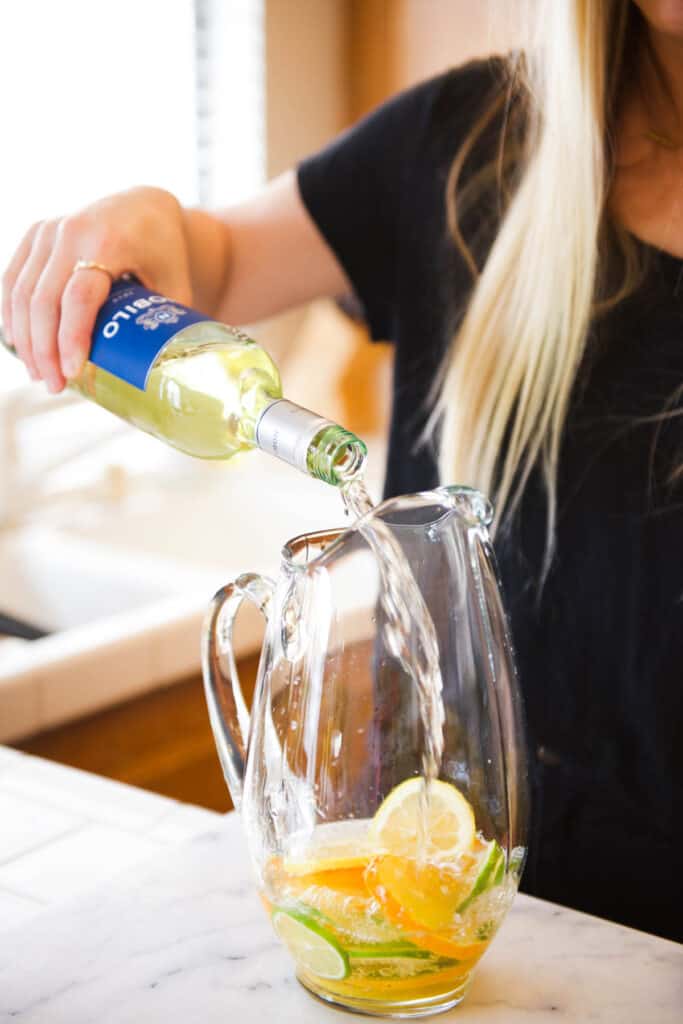 Woman adding white wine into a glass pitcher with sliced citrus to make margarita sangria.