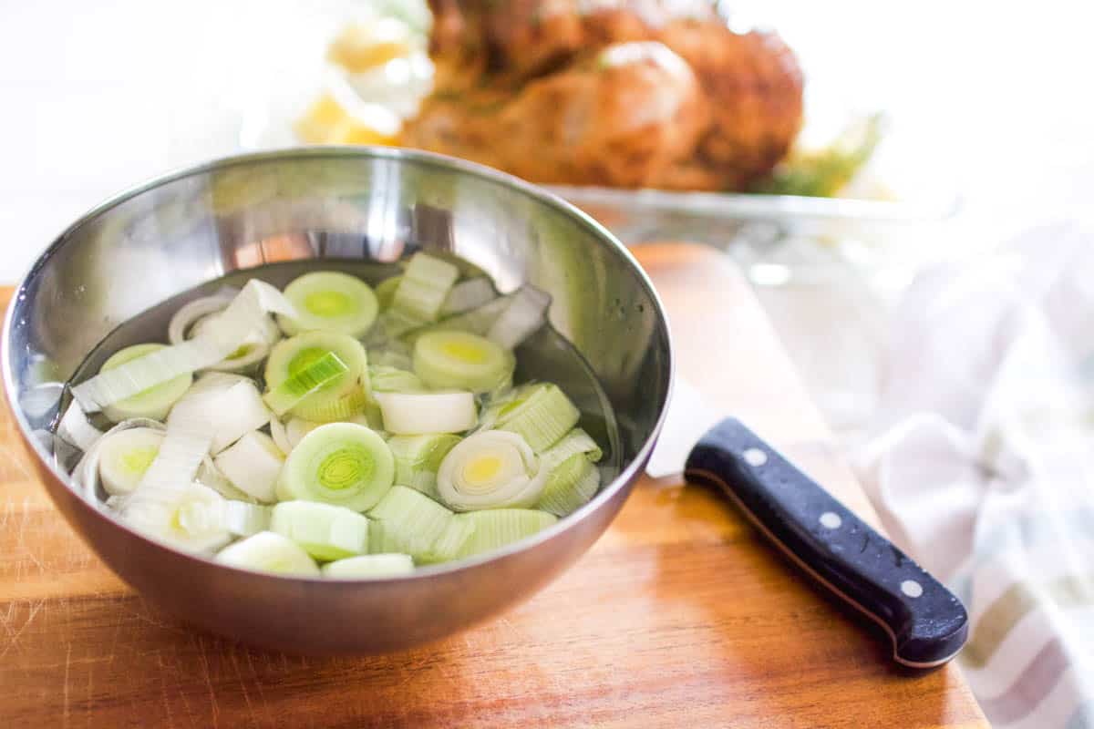 A bowl of sliced leeks on the counter next to a knife and roast chicken.