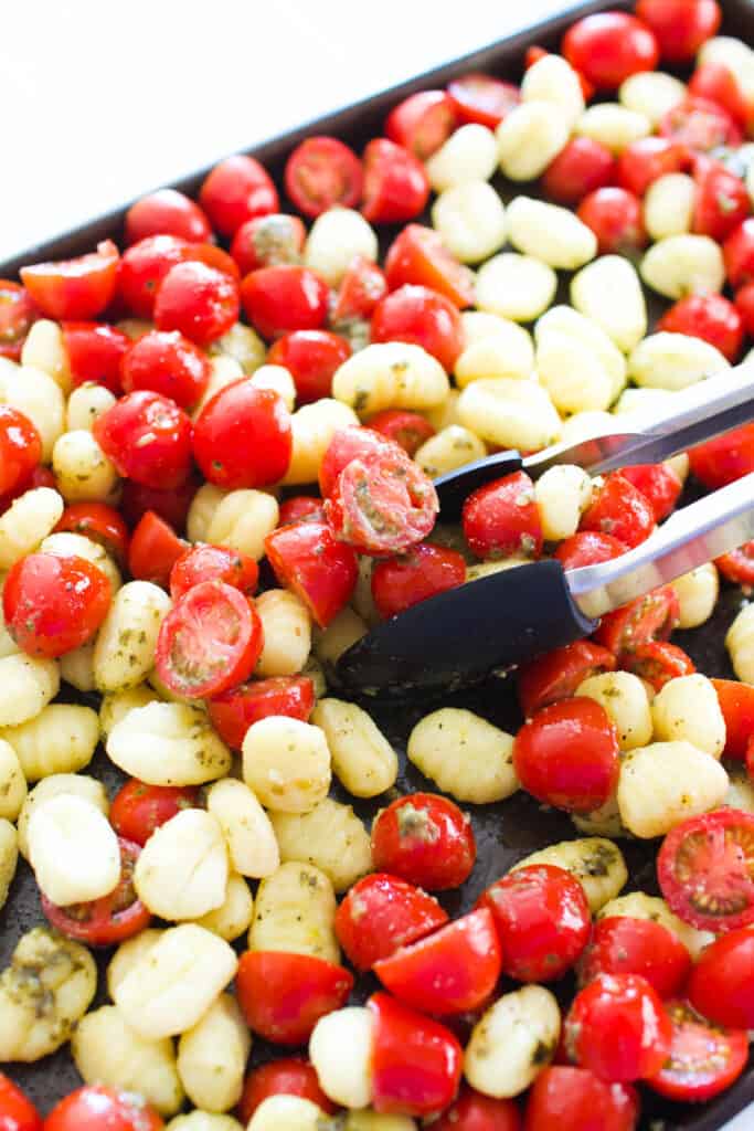 Close up of tongs tossing pesto on tomatoes and gnocchi.