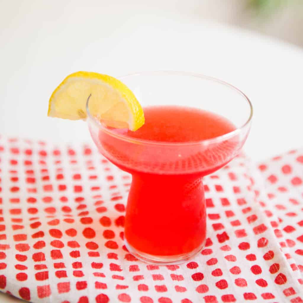 Cocktail glass with a bright pink cocktail and lemon garnish on a pink and white napkin.