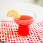 Cocktail glass with a bright pink cocktail and lemon garnish on a pink and white napkin.