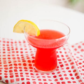 Cocktail glass with a bright pink cocktail and lemon garnish on a pink and white napkin.