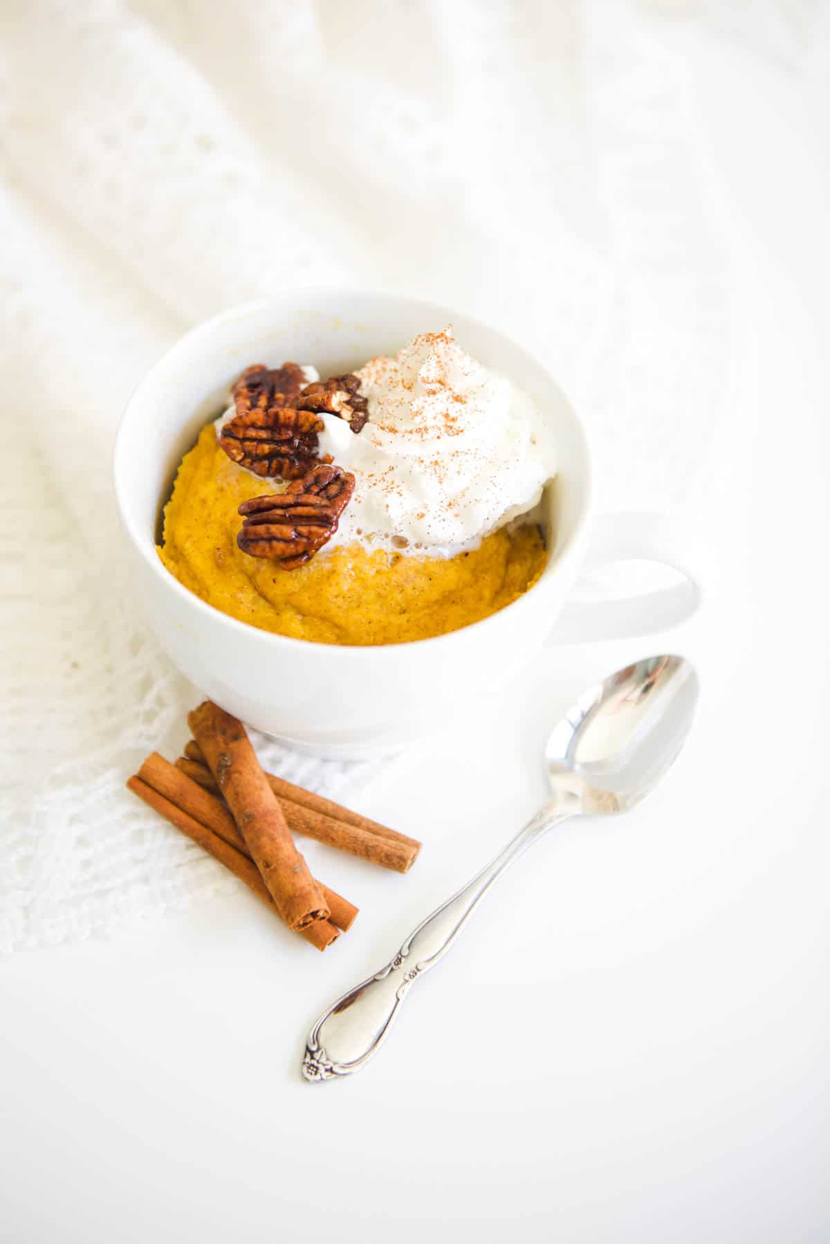 A whip cream topped pumpkin mug cake on a table next to a spoon and cinnamon sticks. 