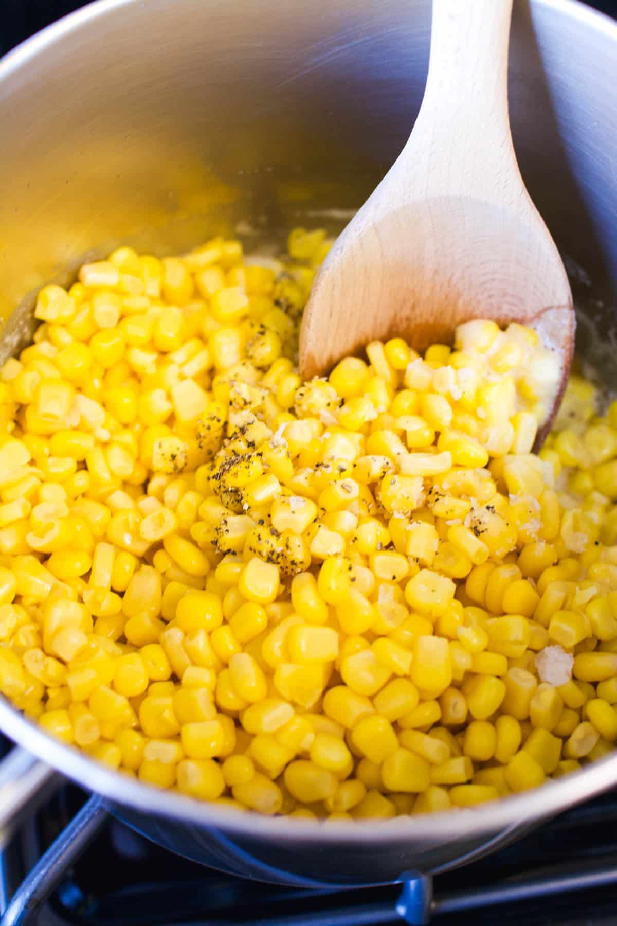 Corn in a saucepan with salt and pepper and a wooden spoon.