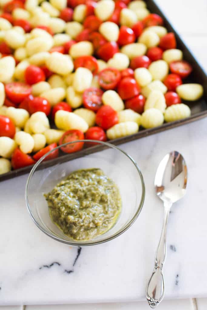 Close up of a small bowl of pesto sauce next to a sheet pan with gnocchi and tomatoes.