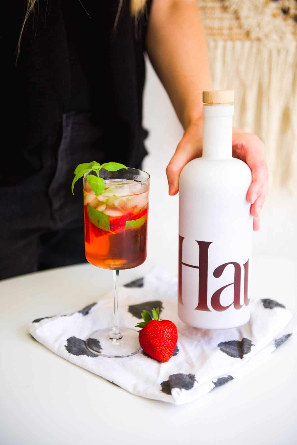 A wine glass holding a cocktail on a table and a woman setting a bottle of Haus on the table next to it.