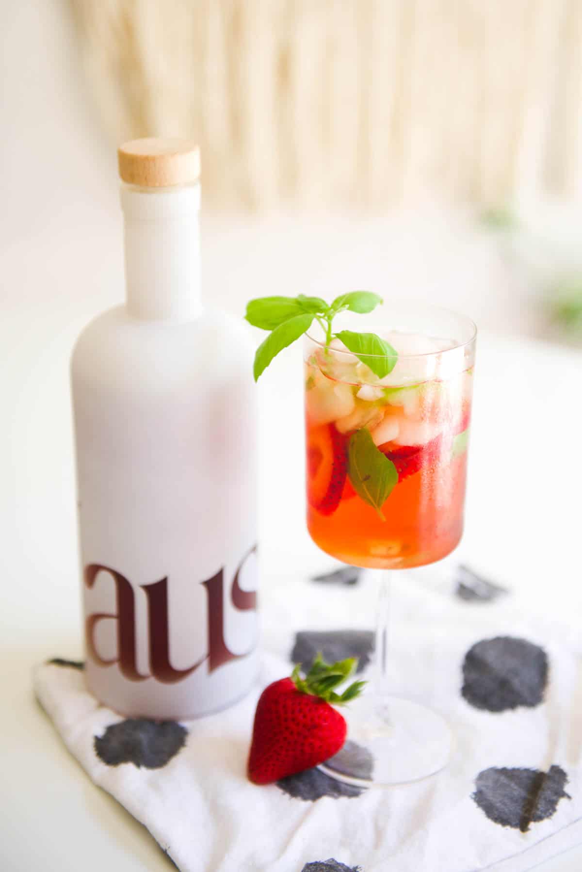 A wine glass on a table with a strawberry basil cocktail next to a bottle of liquor.