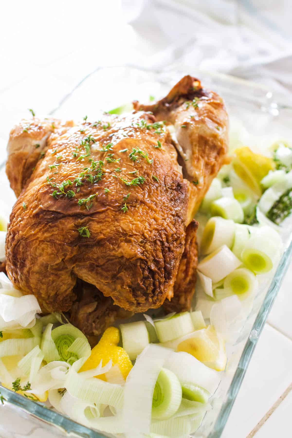 Close up of a roast chicken in a pan with leeks and lemons around it and thyme on top.