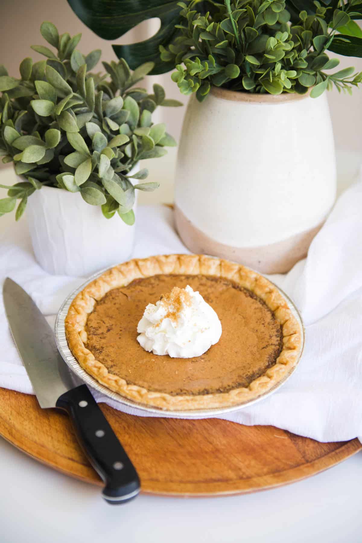 A finished Brown Sugar Pie in a tin on a table next to a knife with a dollop of whipped cream and brown sugar sprinkled on top. 