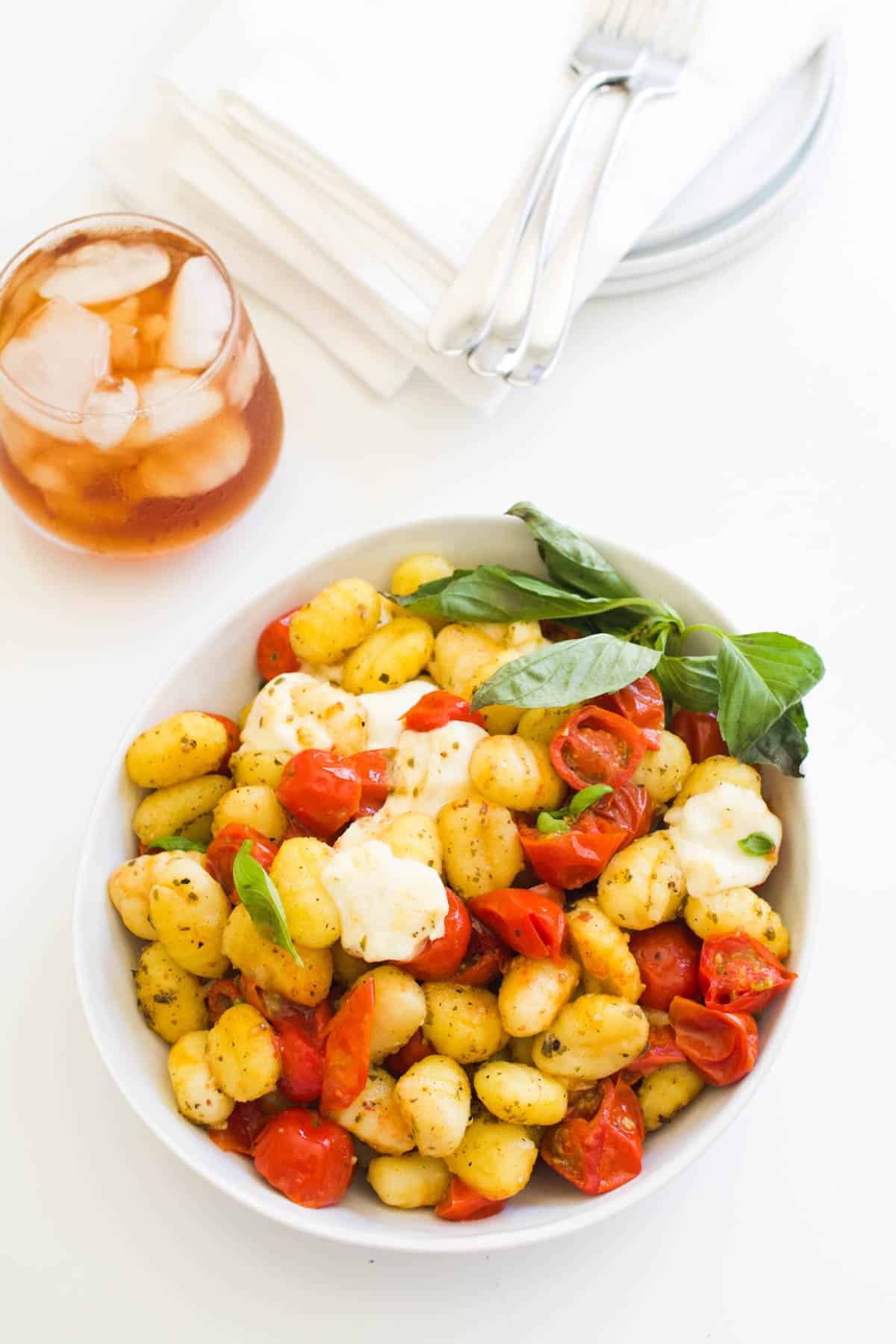 A bowl of gnocchi caprese on a table next to a glass of iced tea.