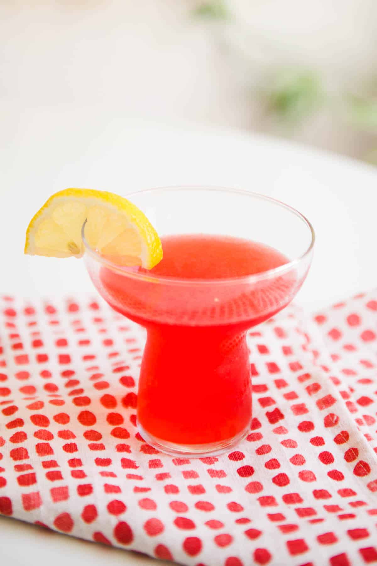 Pink cocktail in a glass with a lemon garnish on a table.