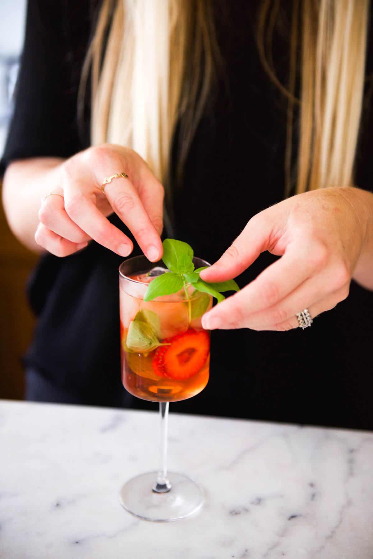 Woman adding a basil garnish to a pink cocktail in a wine glass.