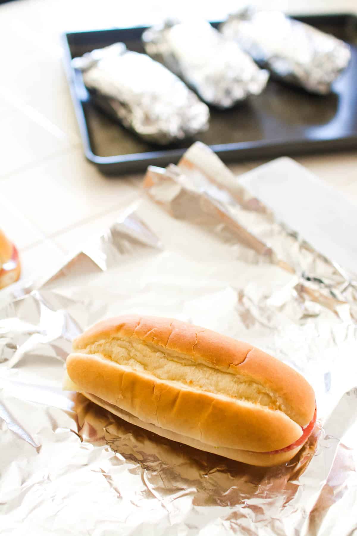 A sandwich on a piece of aluminum foil with more on a tray behind.
