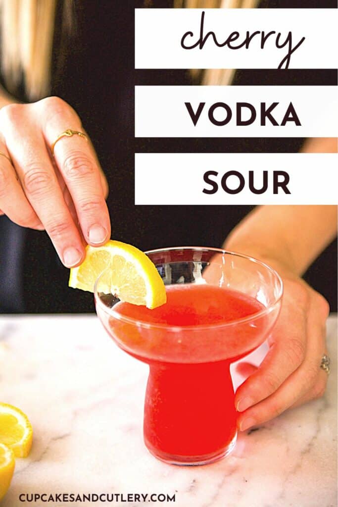 Text - Cherry Vodka Sour with a woman adding a lemon slice to the side of a cocktail glass holding a pink drink.