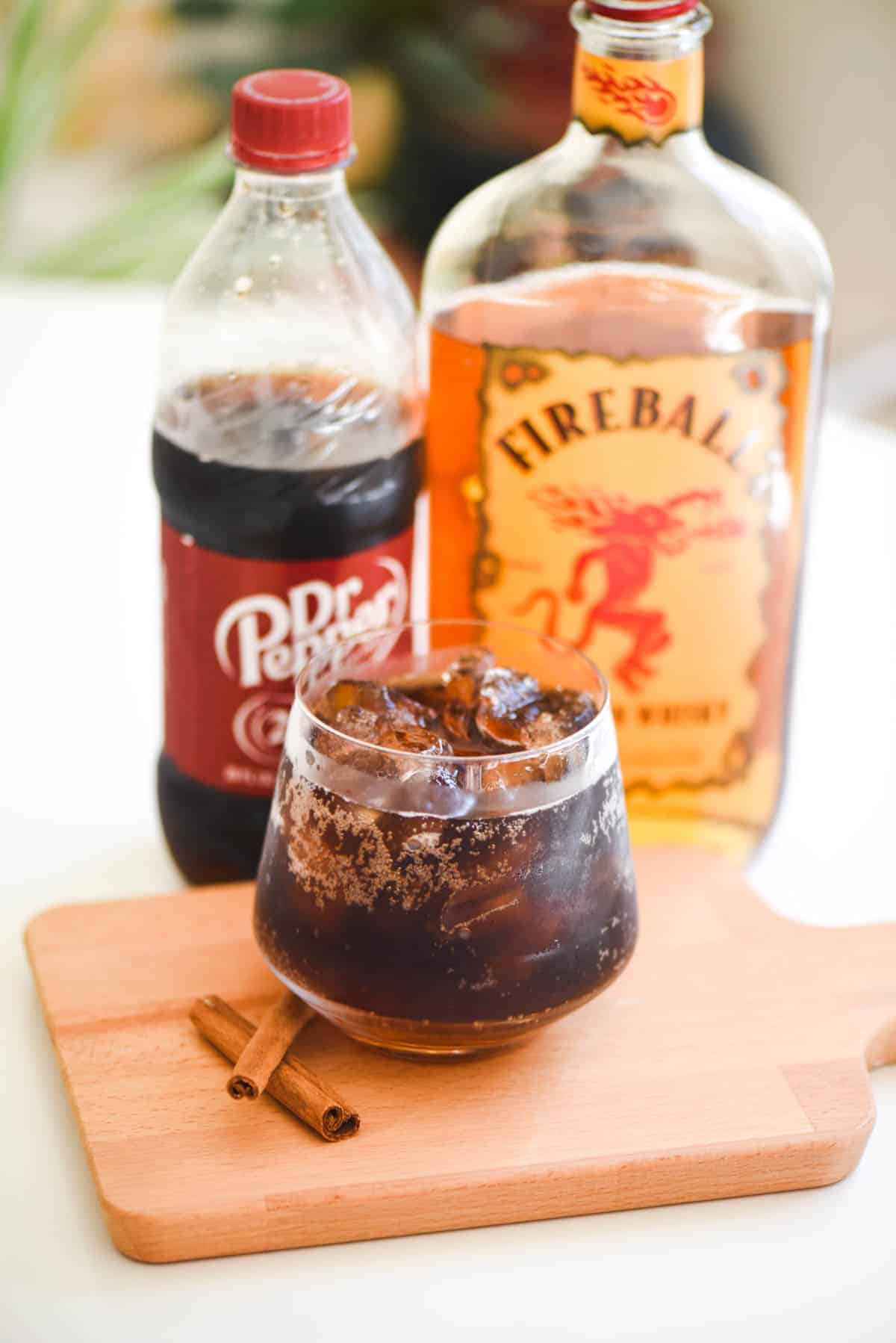 A cocktail in a glass on a counter next to bottles of cinnamon whisky and Dr. Pepper.