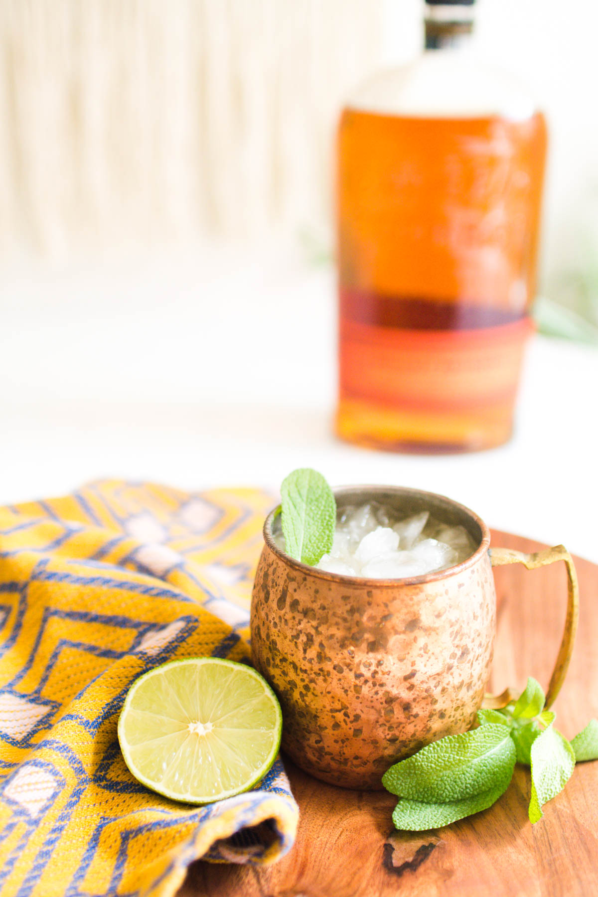 A bottle of bourbon in the backround of a close up shot of a cocktail in a copper mug.