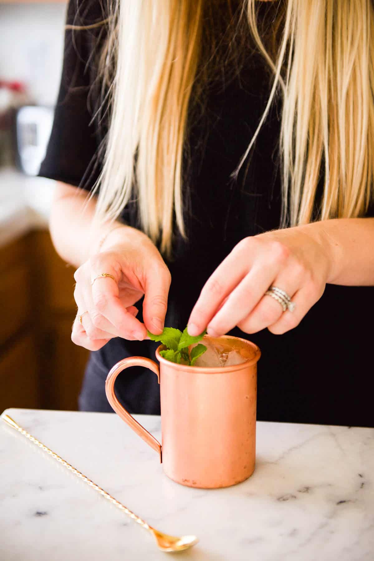 Woman adding a fresh mint garnish to a Moscow Mule mocktail in a copper mug.