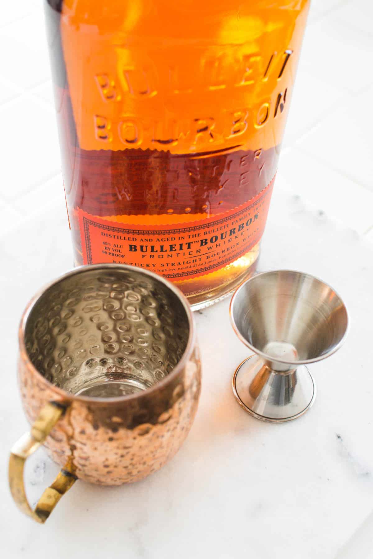 A copper Mule mug next to a jigger and bottle of bourbon on a counter.