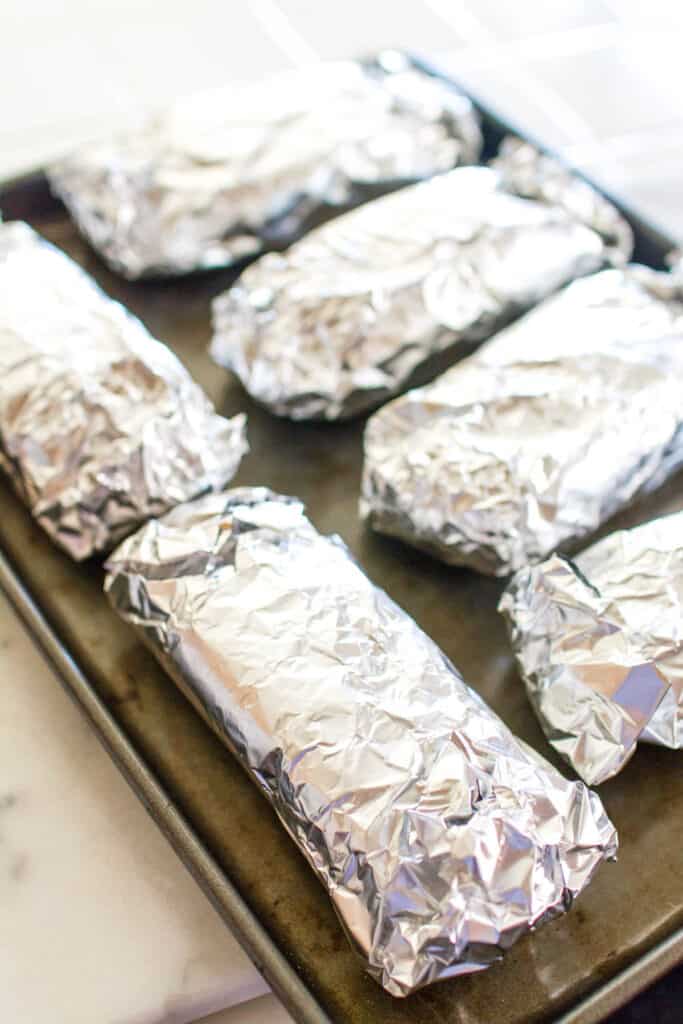 A cookie sheet of sandwiches wrapped in aluminum foil.
